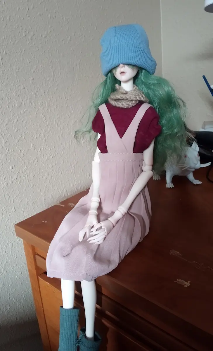 A pale doll sitting on a desk. It has green hair. It's whearing a skirt with suspenders.