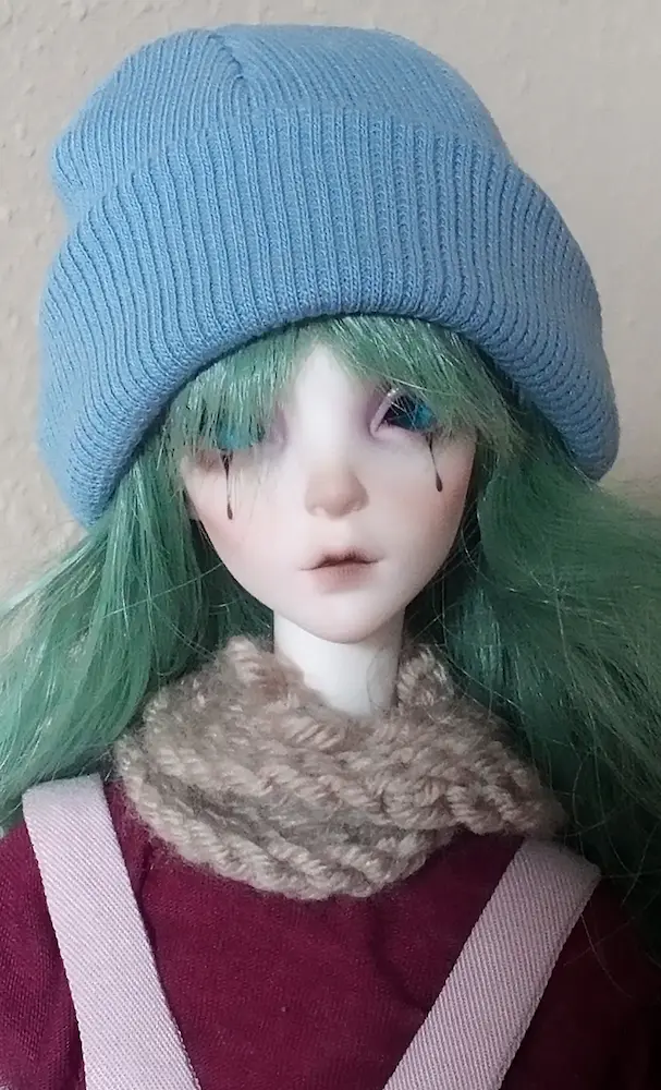 A pale doll with long green hair. It's wearing a beanie and a crochet scarf.